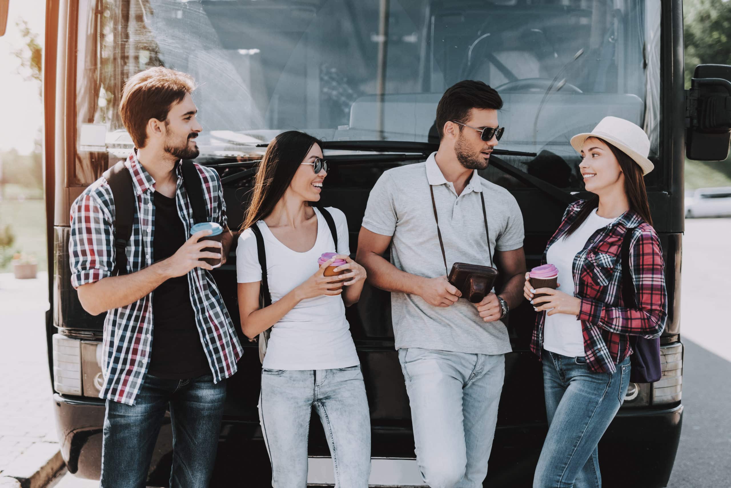 Young People Drinking Coffe in front of Tour Bus. Group of Smiling Friends with Backpacks Standing Together and Talking. Traveling, Tourism and People Concept. Happy Travelers on Summer Vacation